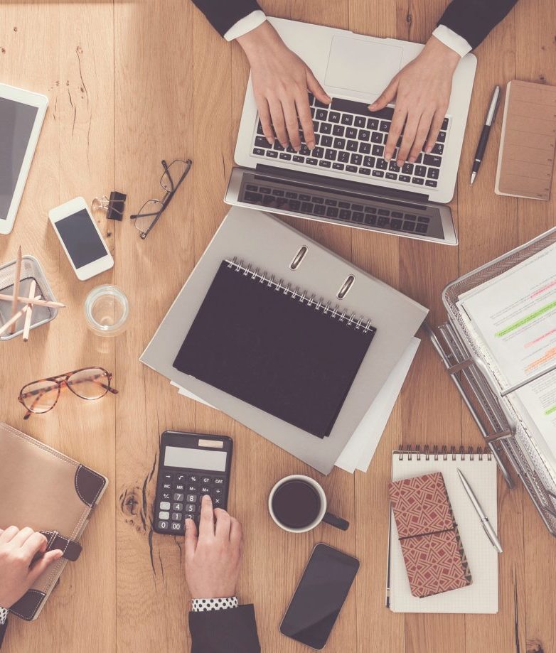 Besprechungstisch aus der Vogelperspektive, darauf sind Laptop, Ordner, Unterlagen, Brillen, Stifte, Notizblöcke und andere Büroutensilien zu sehen. Außerdem zwei Hände die auf dem Laptop tippen und zwei weitere Hände, eine bedient einen Taschenrechner und die andere liegt auf einem Terminplaner